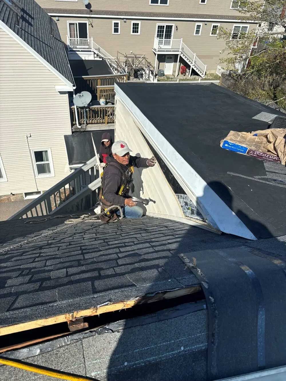 Roofer on a shingled roof, attaching trim to a wall, with a residential building in the background.