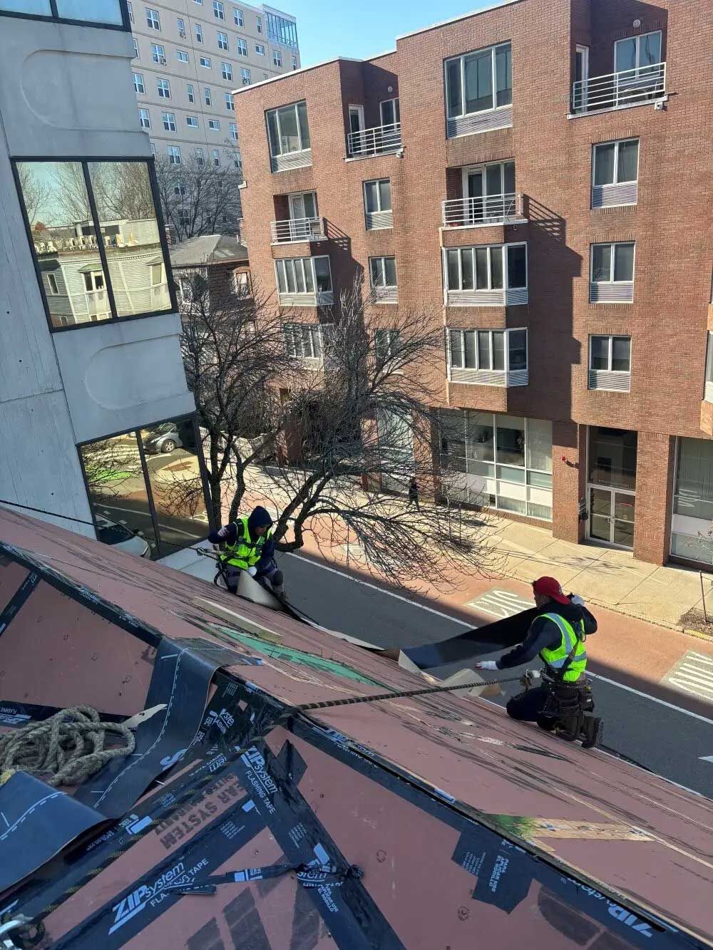 Two roofers in safety vests installing roofing material on a building with a brick facade in the background.