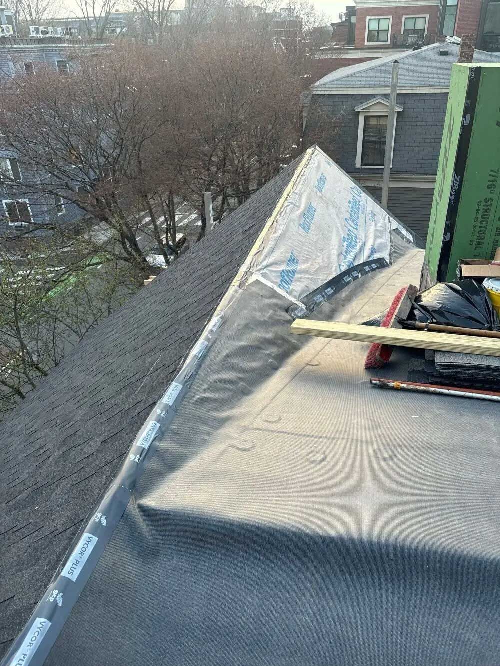 Rooftop view of construction; black roofing material, silver flashing, and wooden tools sit on dark shingles.