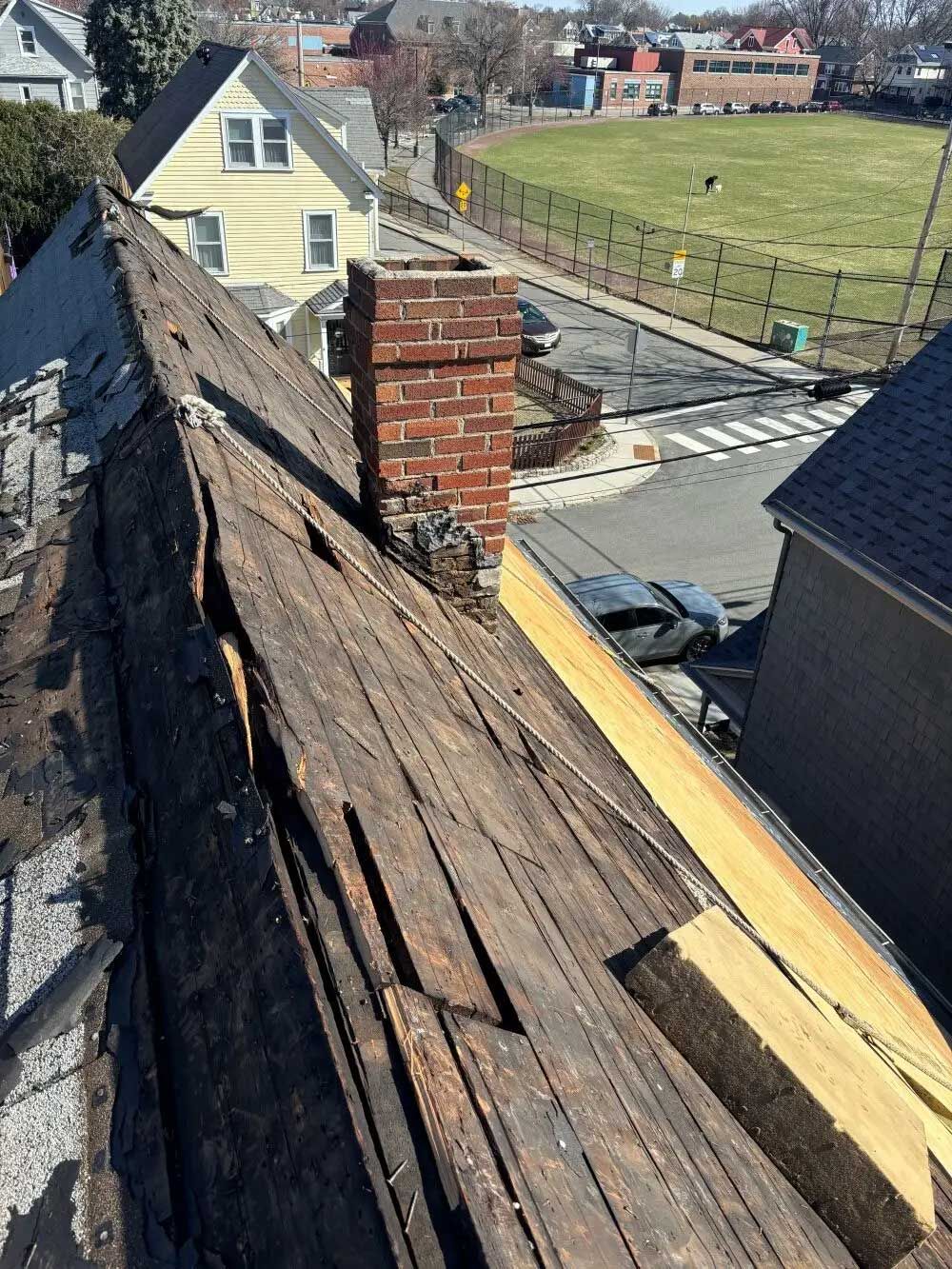 Damaged roof with missing shingles and exposed wood, a brick chimney, and a street scene in the background.