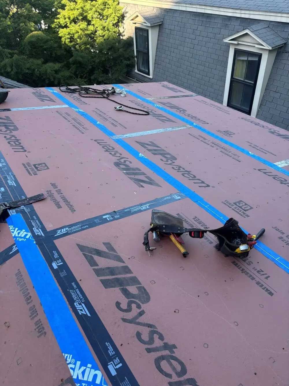 Rooftop with installed Zip System sheathing, tools, and blue tape; light brown and black; sunny day.