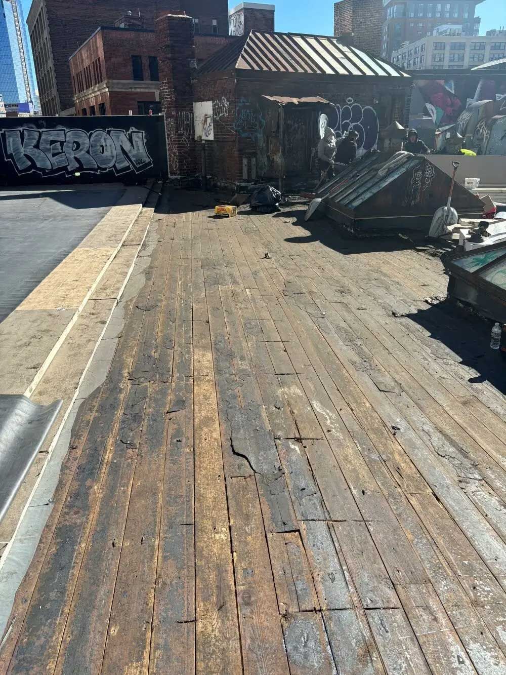 Rooftop with weathered wooden planks and a brick structure. Grayscale graffiti is visible on a nearby wall.