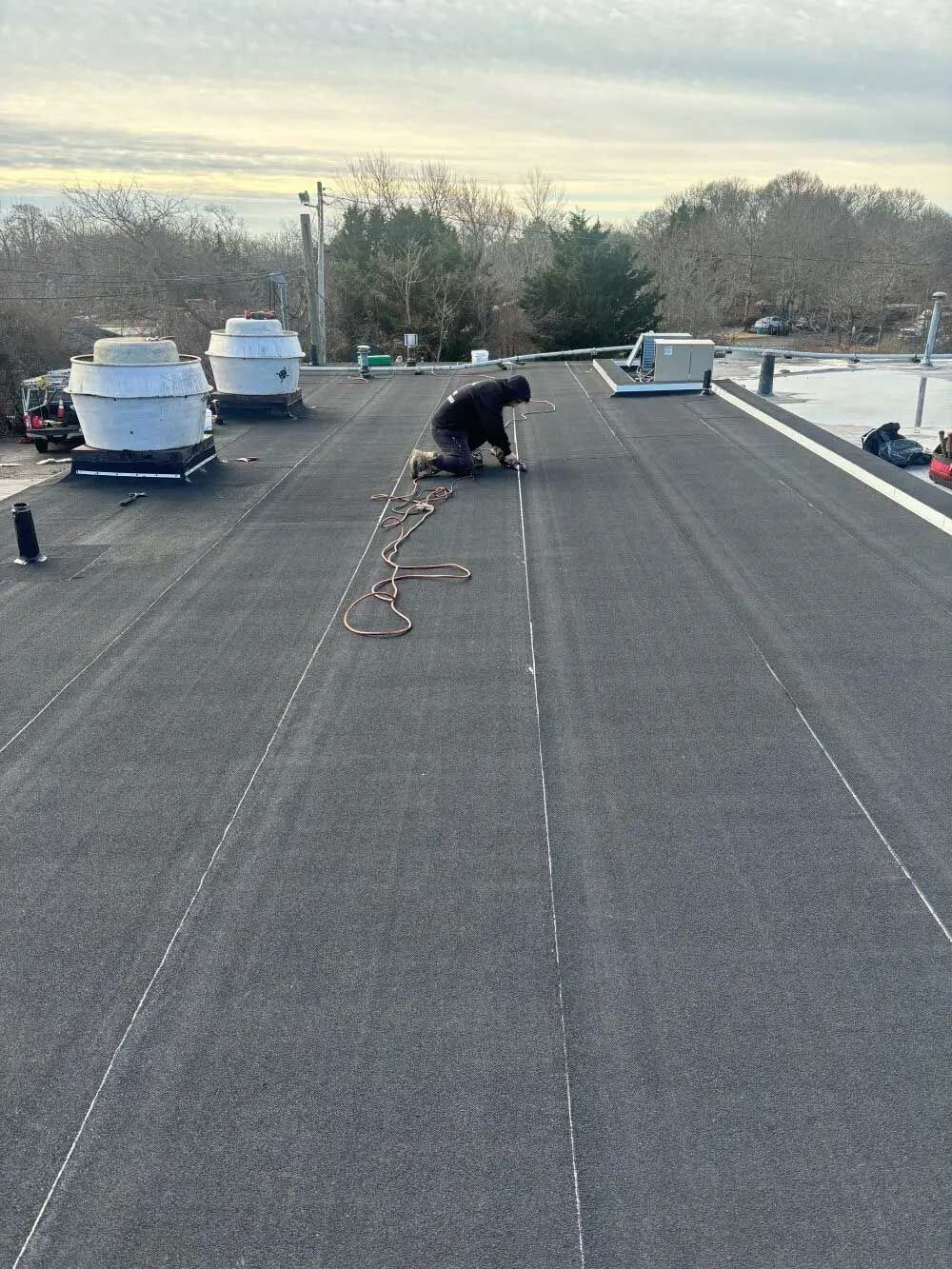 Person kneeling on a flat, asphalt roof, working. Gray sky, trees, and rooftop equipment in background.