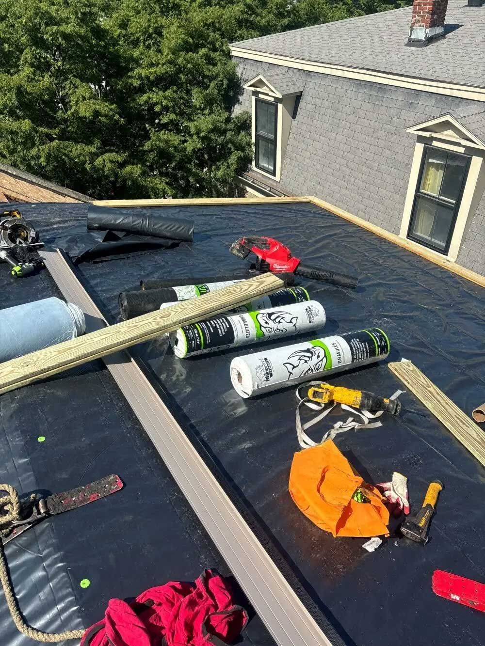 Rooftop with roofing materials: black tarp, rolls, tools, safety gear; gray house and green trees in background.