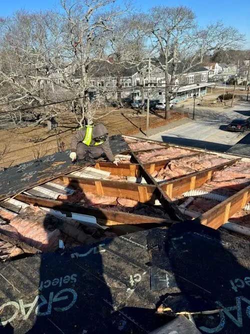 Roofer in safety gear removing shingles from a damaged roof; insulation exposed; sunny outdoor setting.