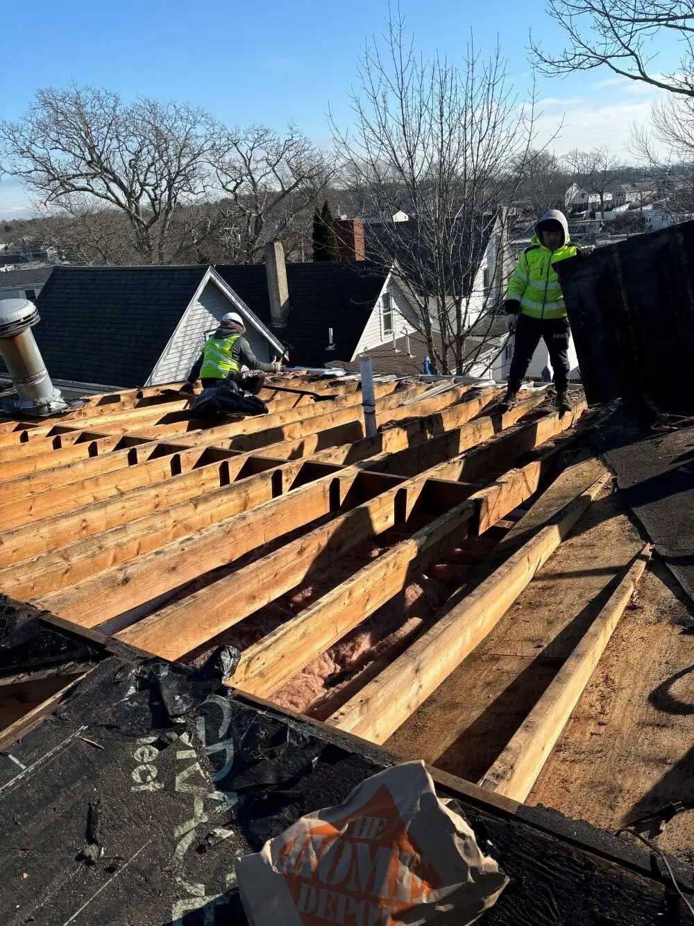 Two roofers in safety vests remove old roof materials from a residential roof on a sunny day.