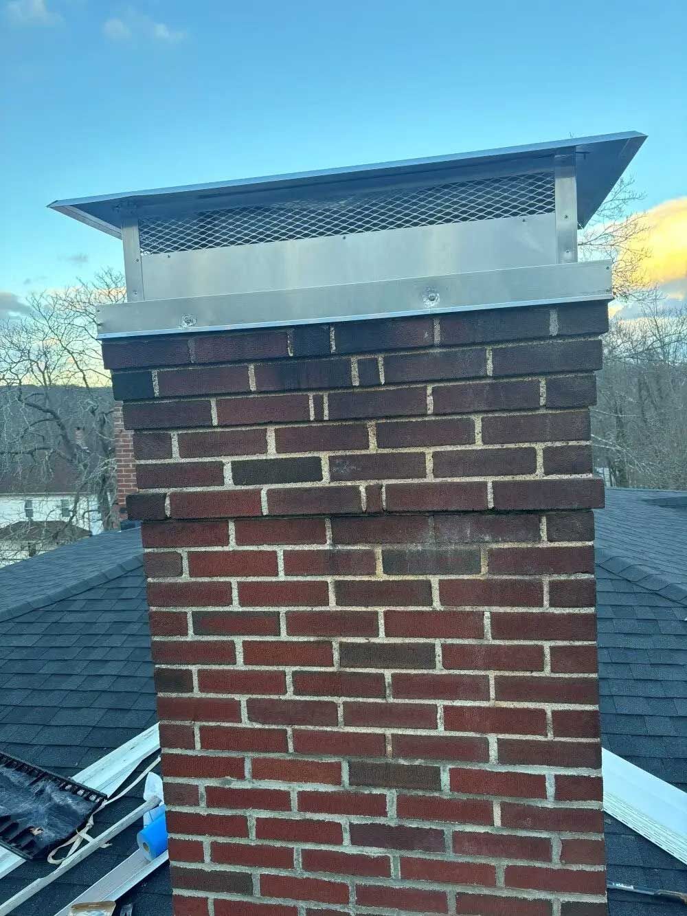 Brick chimney with metal cap on a rooftop under a blue sky.