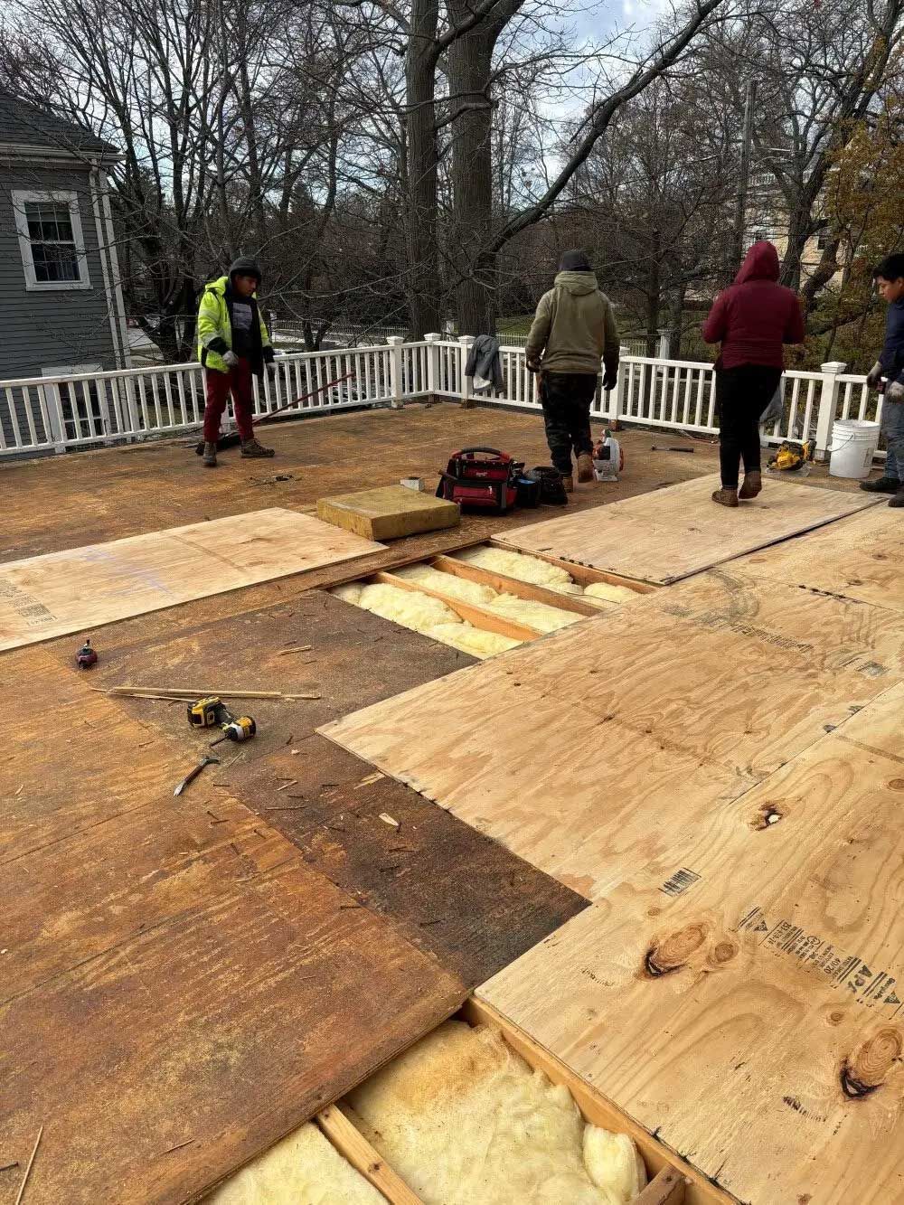 Construction workers replace a wooden deck. Brown plywood and insulation are visible; people are in the background.