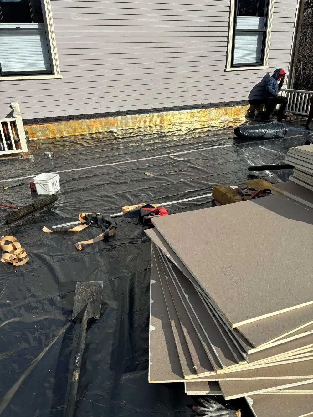 Construction site: Worker installing siding on house. Stack of boards, black tarp, tools, and white railing.