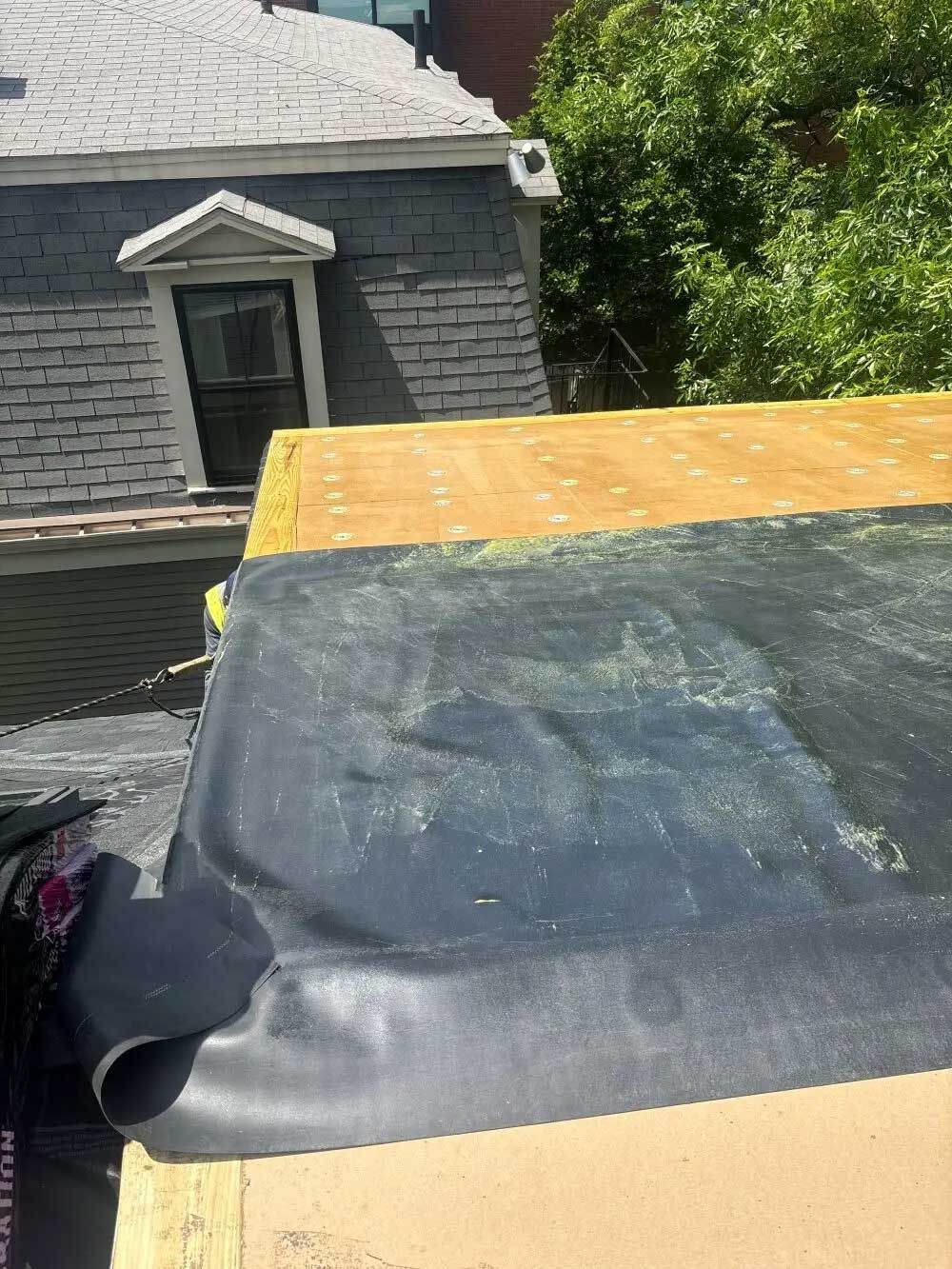 Black roofing material being installed on a flat roof, with a building in the background.