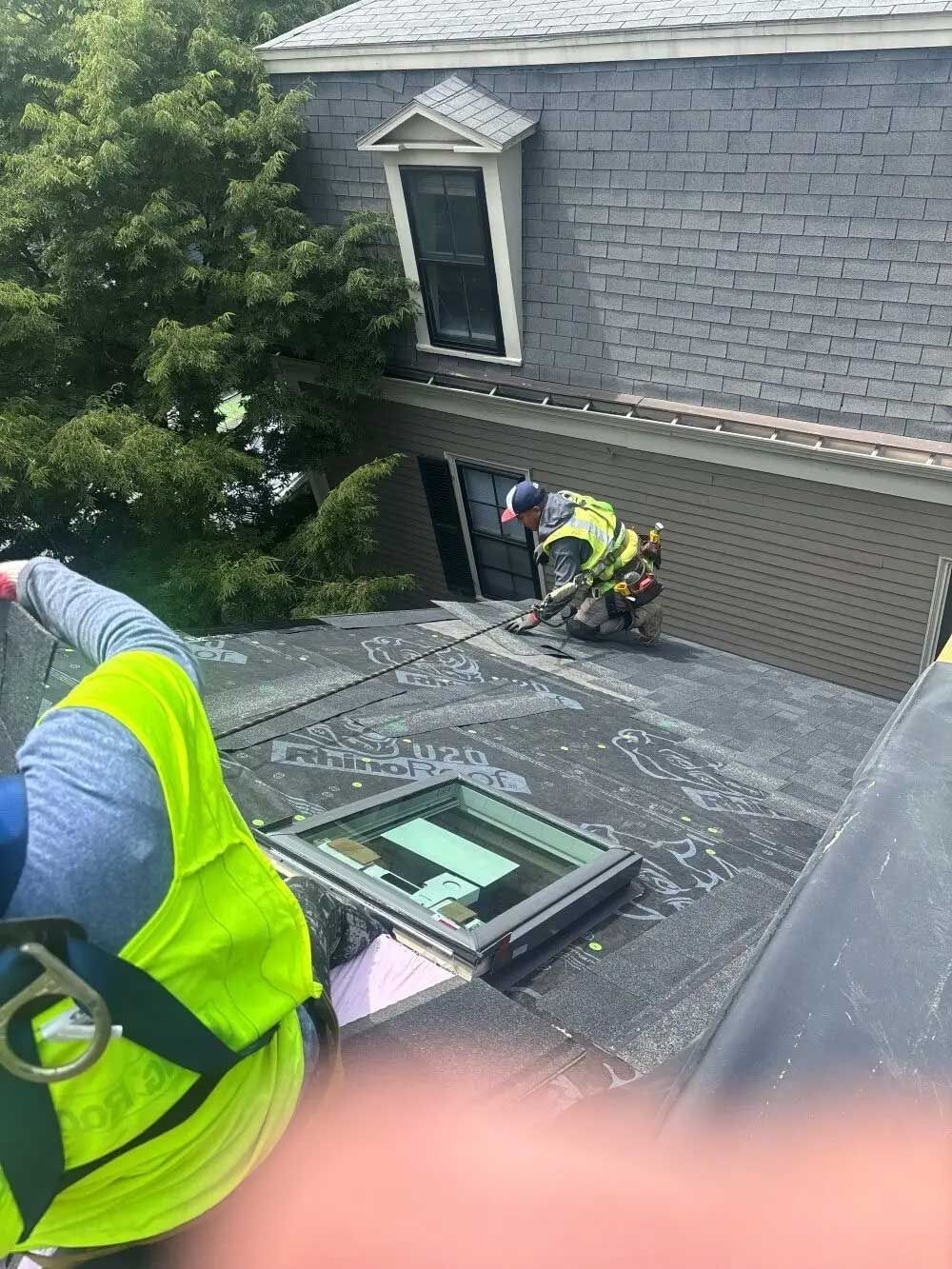 Two construction workers on a roof; one installs flashing, other in vest is partially visible.