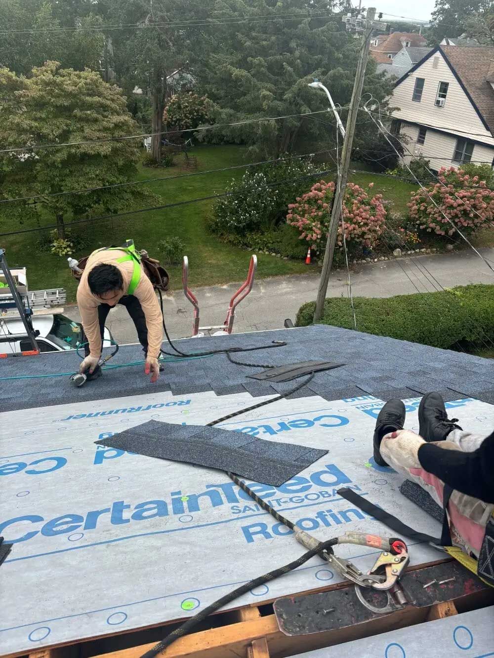 Roofer on a rooftop laying shingles. Safety gear visible, overcast day, residential setting.