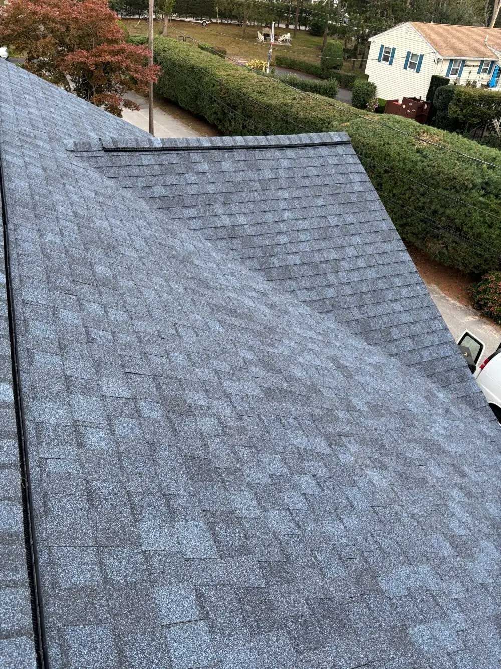 View of a dark gray shingled roof with a house and greenery in the background.