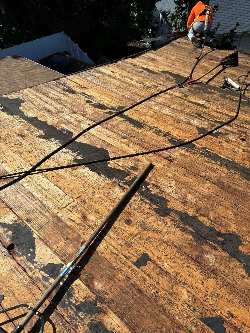 Roofer on weathered roof, removing old material. Wires, dark streaks across brown surface, sunny outdoor setting.