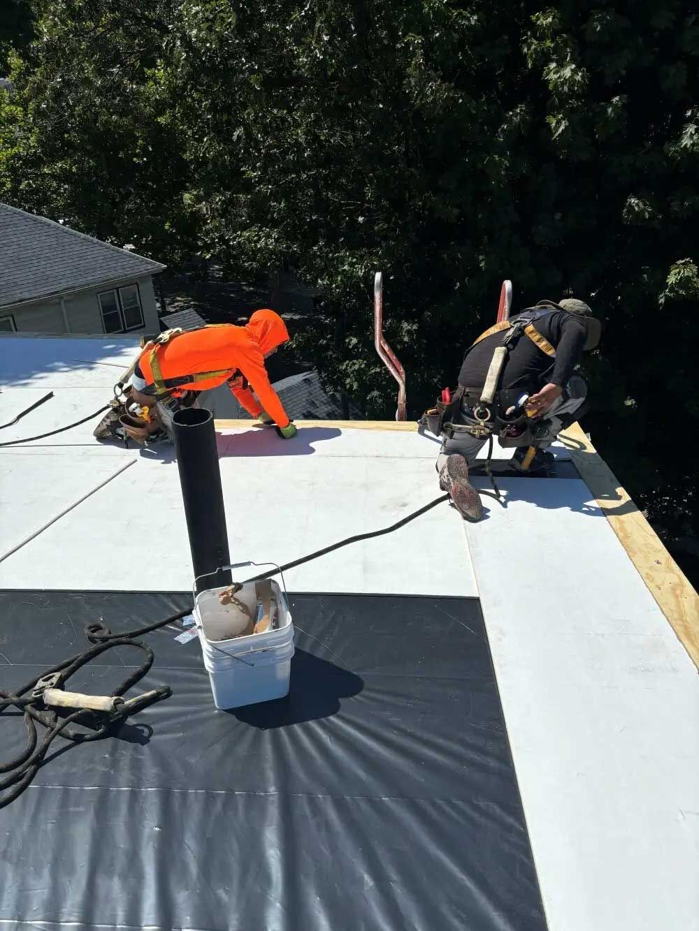 Two roofers in safety harnesses on a white roof, one in orange, the other in black, working under a sunny sky.