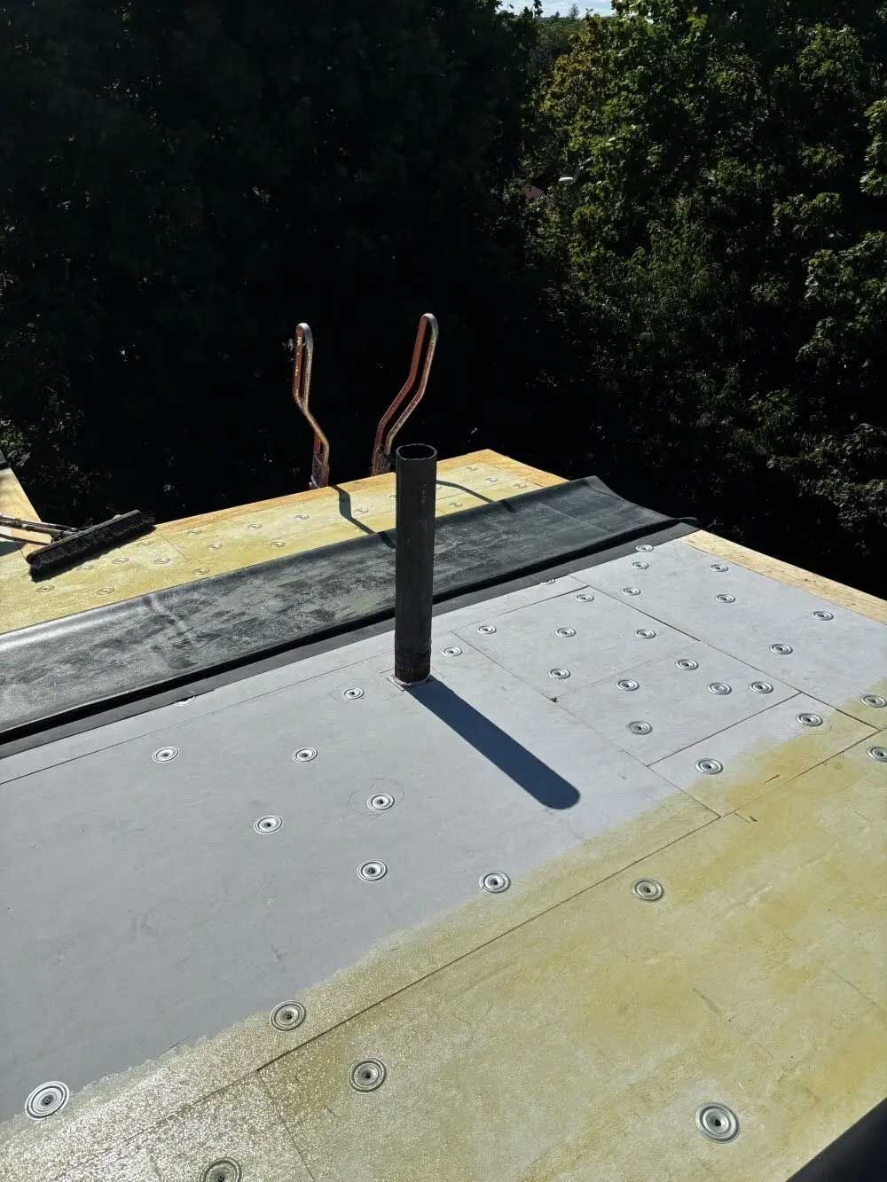 Black pipe and wires on a flat roof, sunlight casting shadows, surrounded by insulation and trees.