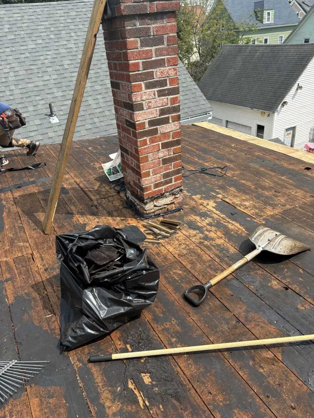 A person repairs a roof near a brick chimney, with a black trash bag and shovel on the wood surface.