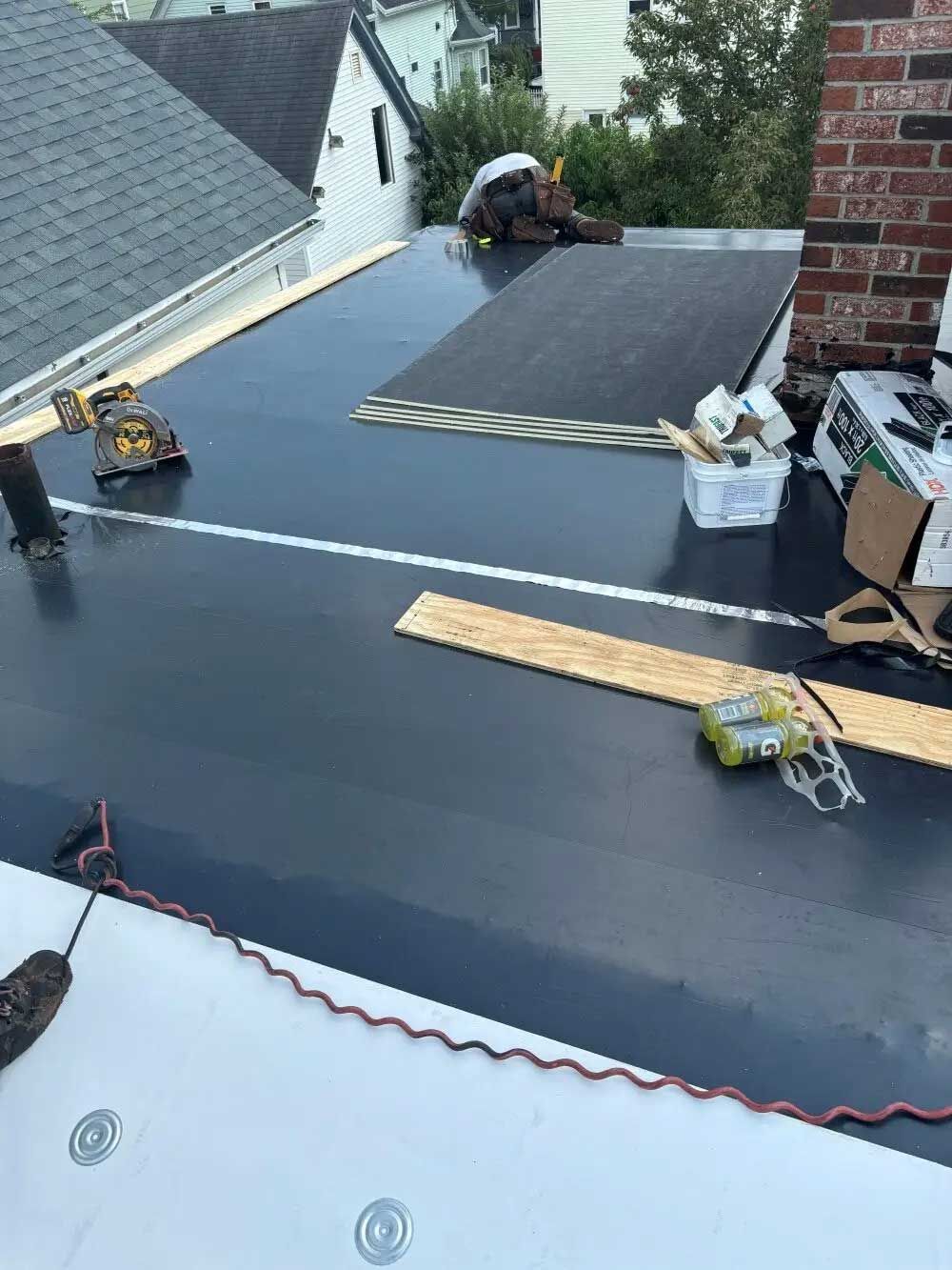 Person on a flat roof installing black roofing material near a chimney and tools.