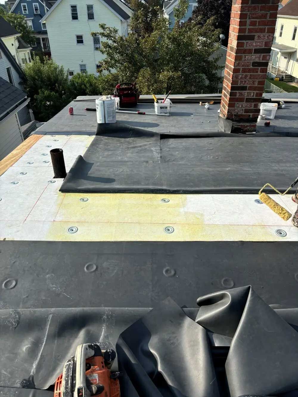 Roof construction: Black roofing material being laid on a flat roof, near a brick chimney.