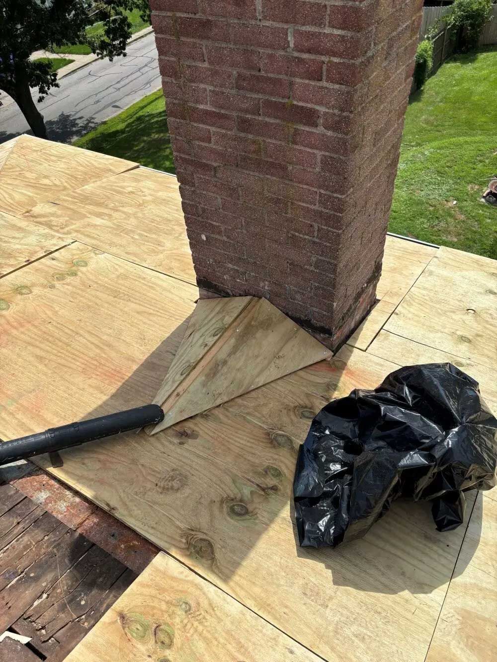 Wooden roof with brick chimney; plywood, black bag, and blower present on the roof.
