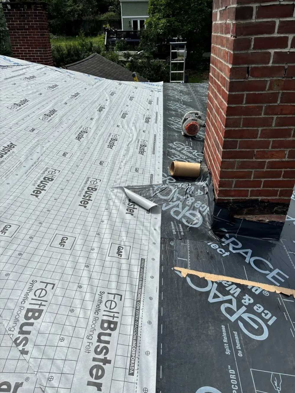Roof under construction; black ice and water shield next to brick chimney, roll of felt, and a gray felt sheet.