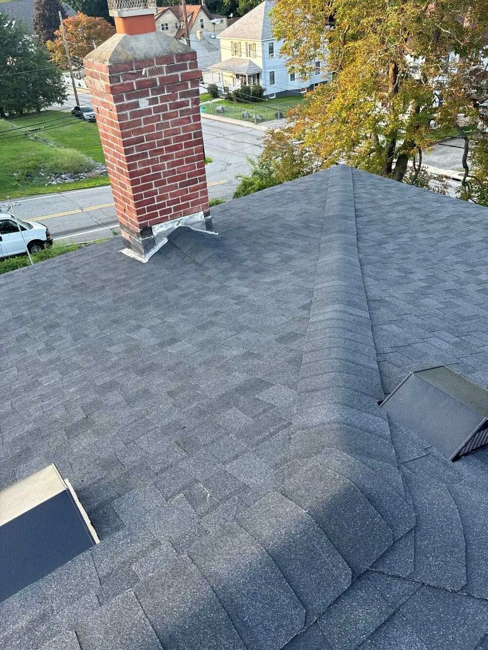 Rooftop with a brick chimney. Grey asphalt shingles. Autumn trees and houses in the background.
