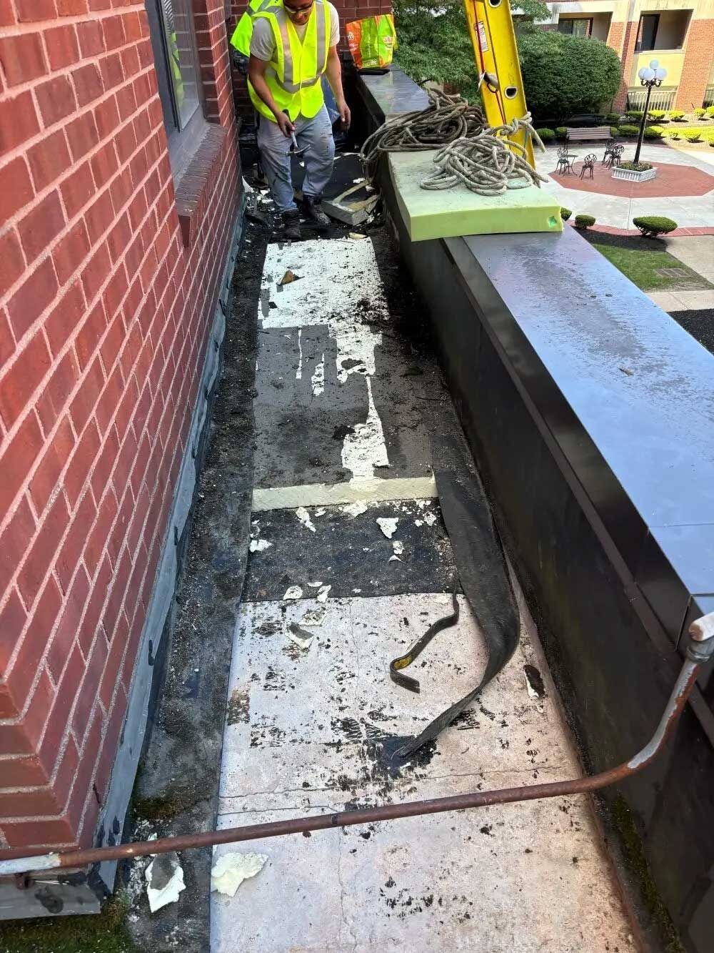 Workers in safety vests repair a narrow roof ledge along a brick building.