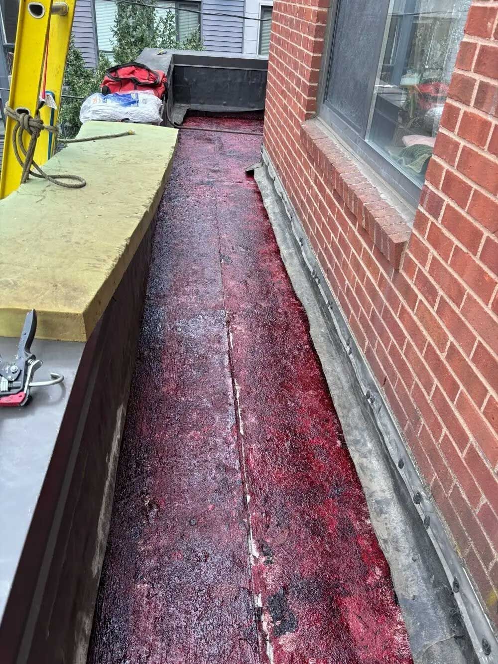 Narrow rooftop walkway with red sealant against a red brick building. Ladder and materials visible.