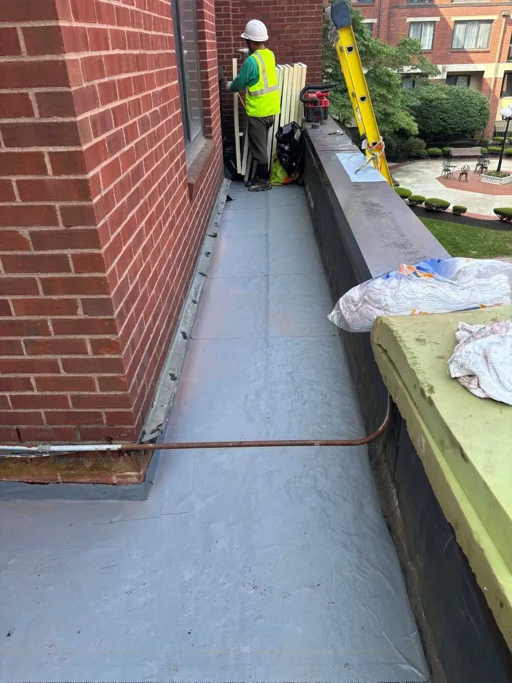 A worker on a rooftop in a reflective vest, prepping a section of gray surface near a brick wall.