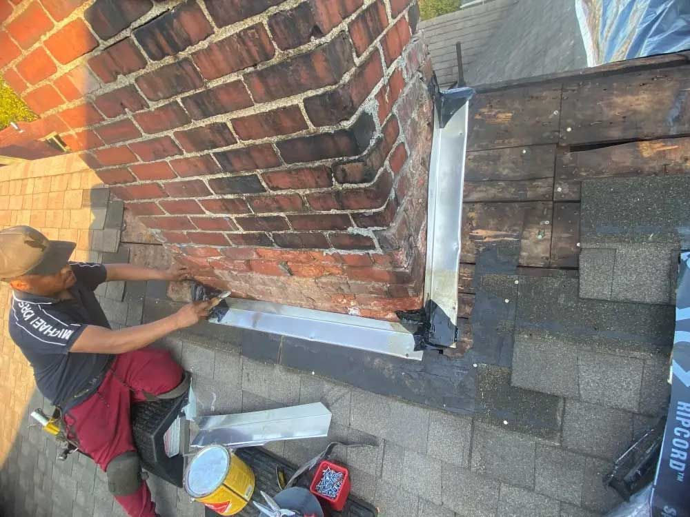 Person on a roof repairs chimney flashing with tools, near shingles.