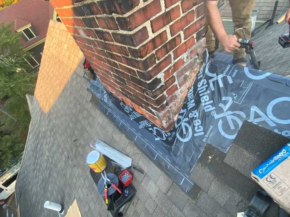 Roofer installing flashing around a brick chimney on a shingled roof.