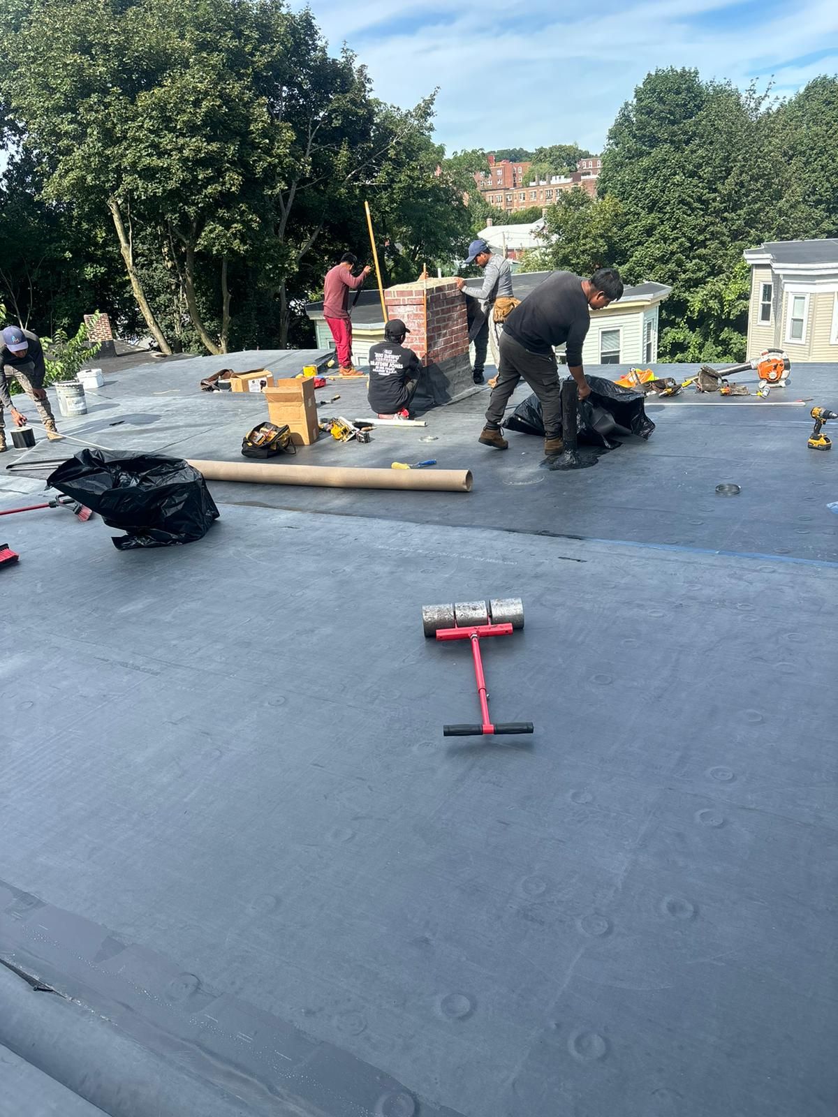Roofers working on a flat roof, tools and materials scattered. Blue sky, trees in the background.