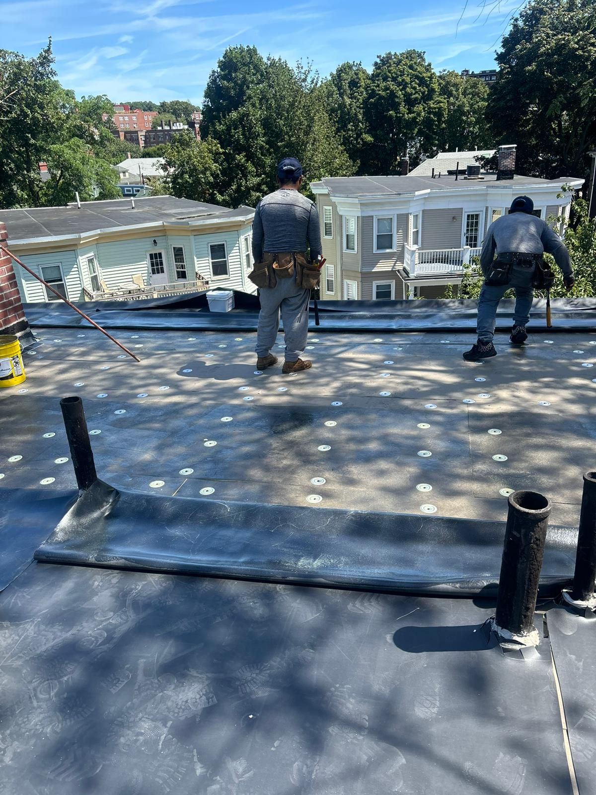Two roofers on a flat roof, one facing away, the other working. Blue sky, rooftops in the background.