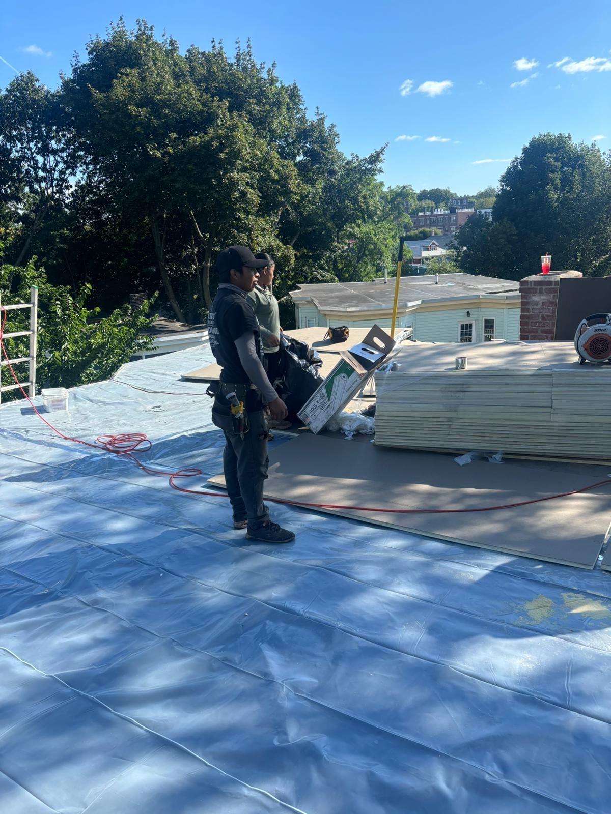 Two workers on a rooftop, one holding a saw, blue tarp, stacks of material, sunny day.