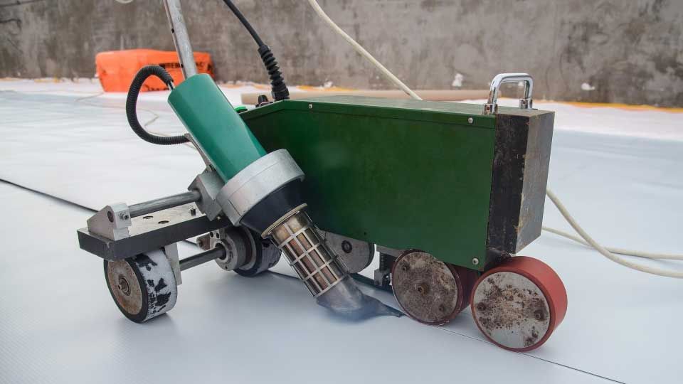 Green welding machine sealing white roofing material on a construction site.