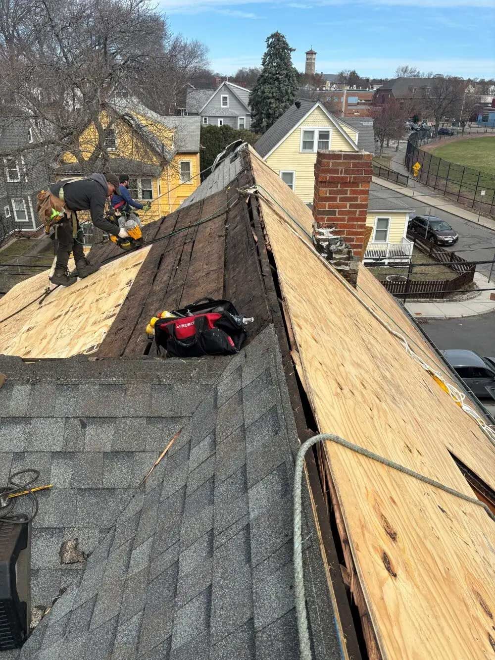 Roofing workers repairing a steep shingled roof with exposed plywood and tools near a street.