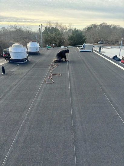 Person kneels on a rooftop, working near vents and loose cables at sunset.