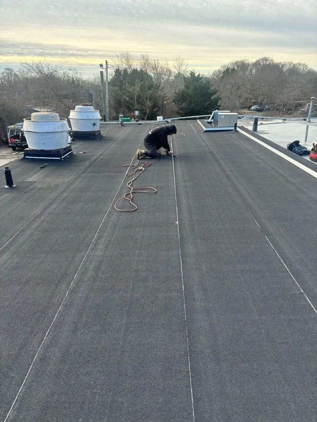 Person kneels on a rooftop, working near vents and loose cables at sunset.