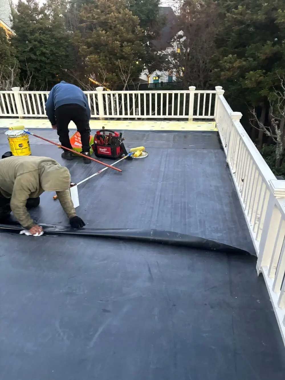 Two workers installing black roofing on a deck with white railings, one rolling out material and one crouching nearby