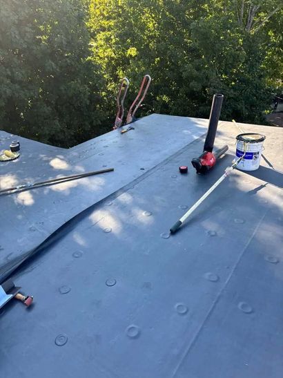 Roof under repair with black membrane, tools, and a paint can on a sloped rooftop by trees