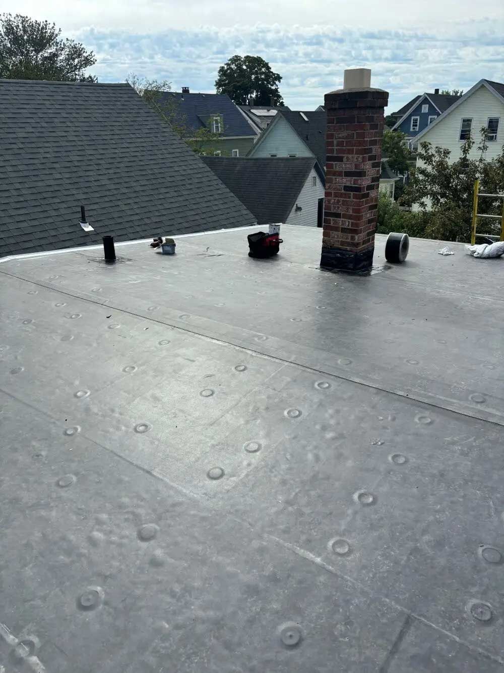 Flat gray rooftop with a brick chimney and vents, overlooking houses under a cloudy sky
