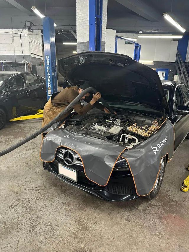 A mechanic is cleaning the engine in the auto shop. The black car has an open hood, covered with a gray shield.
