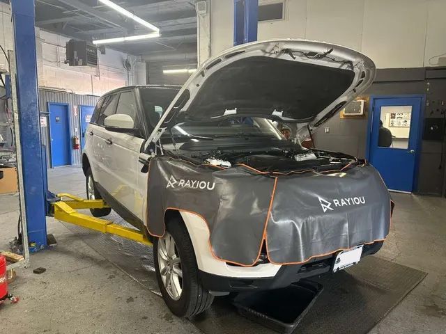 White SUV raised in a repair shop with hood open, covered by a protective pad.