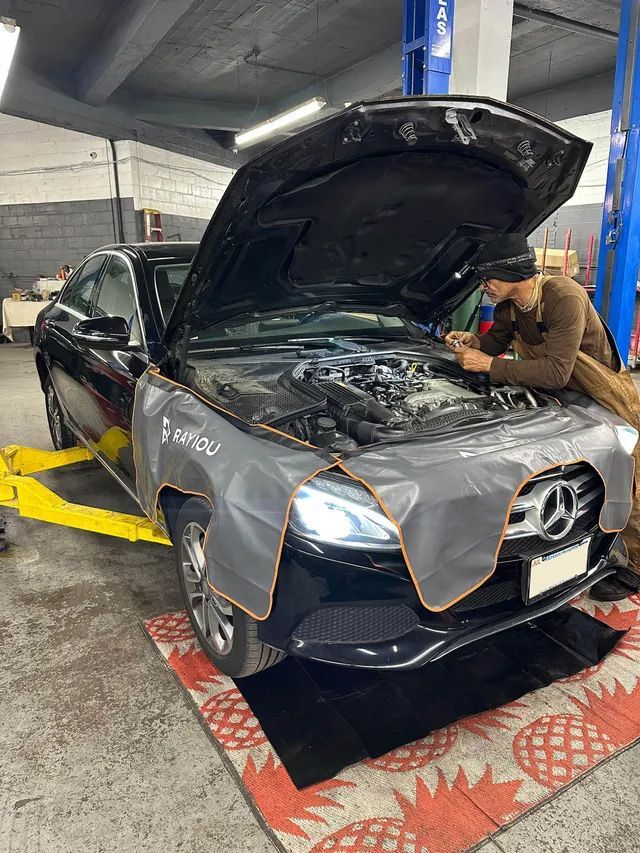 Mechanic working on a black Mercedes with the hood open in a repair shop.