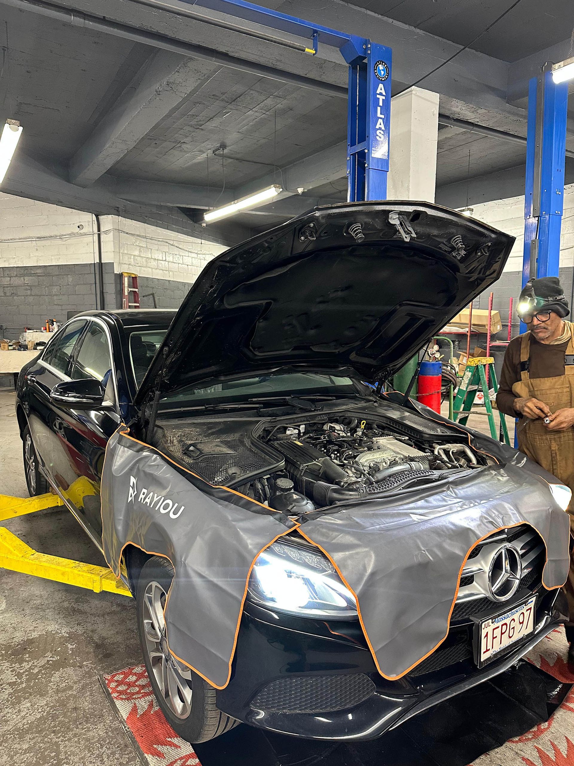 Black Mercedes with hood open on a lift in a garage, a mechanic standing beside it.