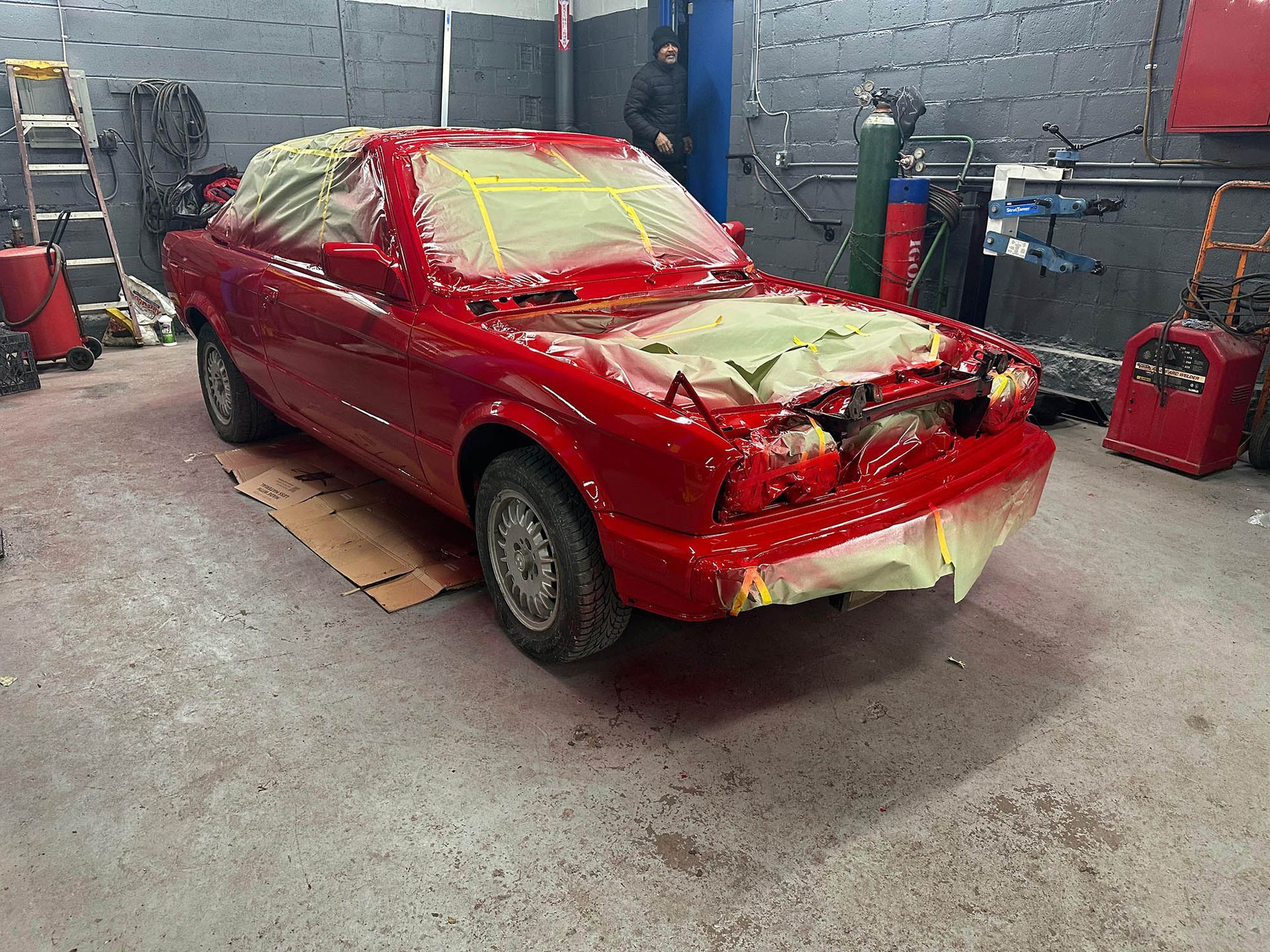 Red car in a garage being painted; masked areas, cardboard on the floor, equipment in the background.
