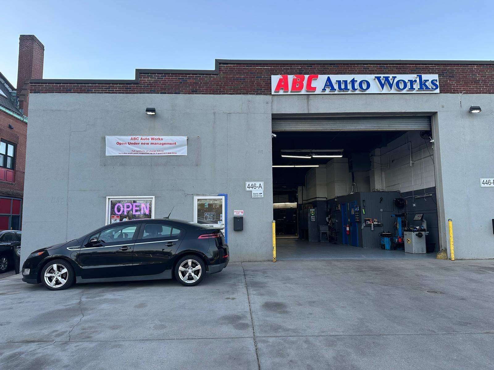 A black car parked outside of ABC Auto Works, a garage with its large door open, under a blue sky.