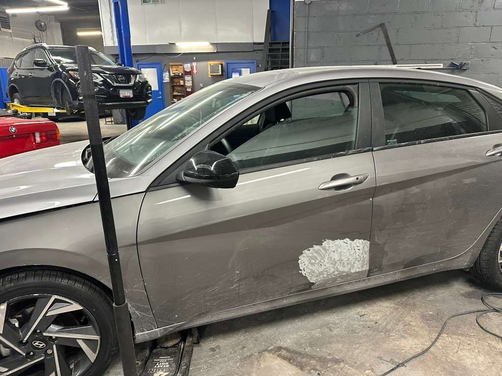 Gray car in a repair shop with a damaged side panel, tire jacked up.
