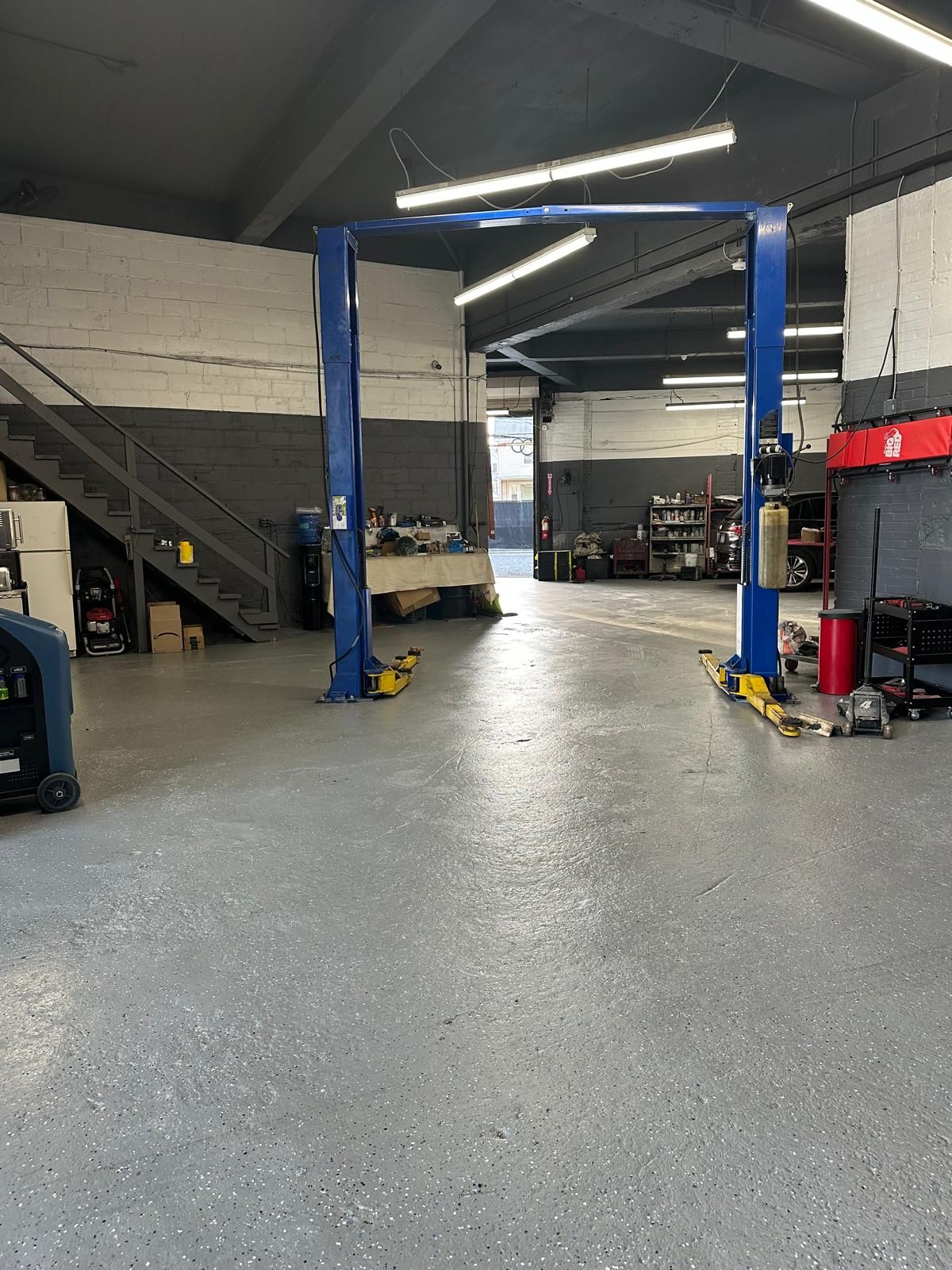 Blue car lift in a gray-floored auto repair shop. Fluorescent lights and stairs visible.