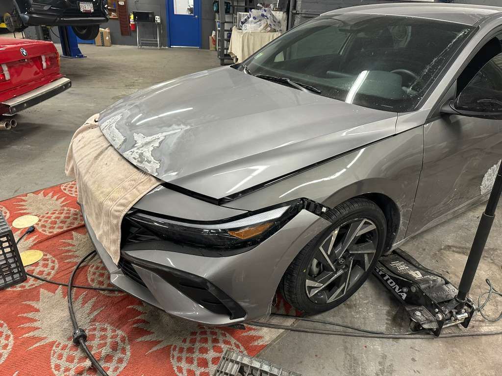A gray sedan being worked on in a garage. The hood is covered with a towel. The front tire is lifted by a jack.